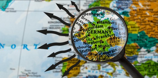 A black magnifying glass is held over a colourful map, focussed on Germany and the rest of Europe