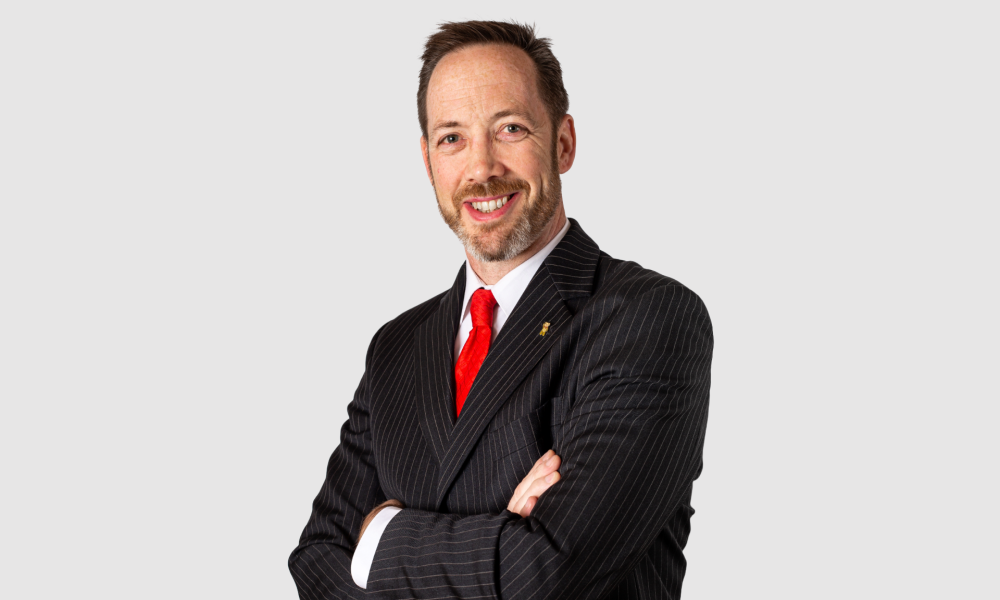 Adam Parry, Managing Director of HELU UK smiles with his arms crossed while wearing a black suit, white shirt and red tie stood against a grey background.