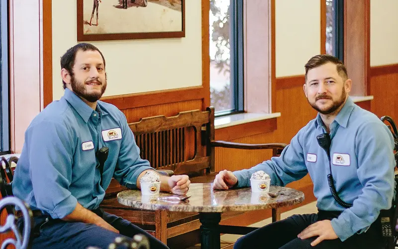 two men sitting at a table, laughing into the camera, with ice cream in a cup in front of them