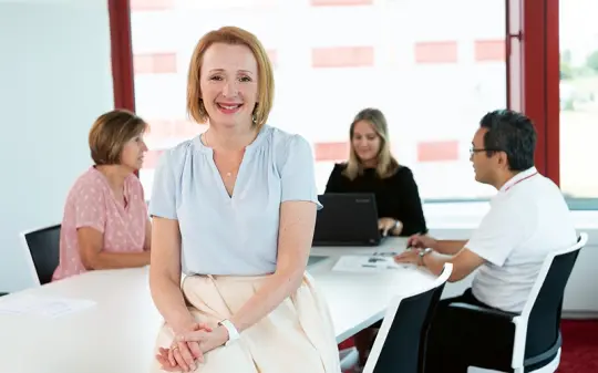 Katja Lägeler leaning against the desk at which a team is working.