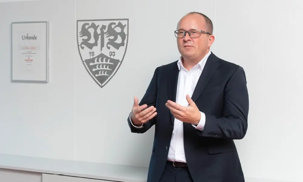 Markus Schöck steht in seinem Büro und im Hintergrund sieht man ein VfB Logo